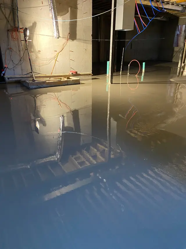 Self-leveling concrete being poured across an unfinished basement floor with framing and plumbing roughed in