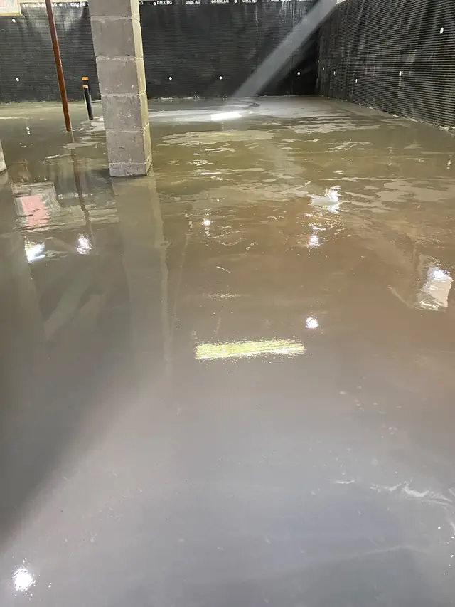 Glossy wet self-leveling concrete on a basement floor beside poured concrete foundation walls