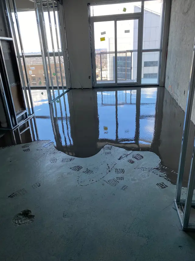 Self-leveling concrete flowing across a primed slab in a condo unit, showing the wet pour line